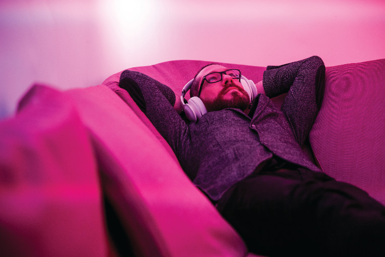 Man wearing over ear headphones lying on his back on a sofa.